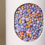 Close-up of colorful coated wildflower seed pellets inside the window of Fleuryx California Bloom Native Wildflower Mix shaker tube, with each pellet color-matched to the flower species it contains.